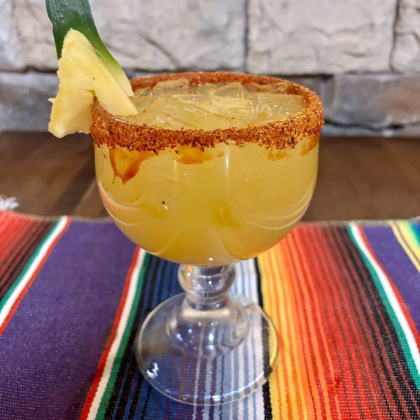 A pineapple margarita with a chili-lime rim and a pineapple wedge, on a colorful striped placemat.