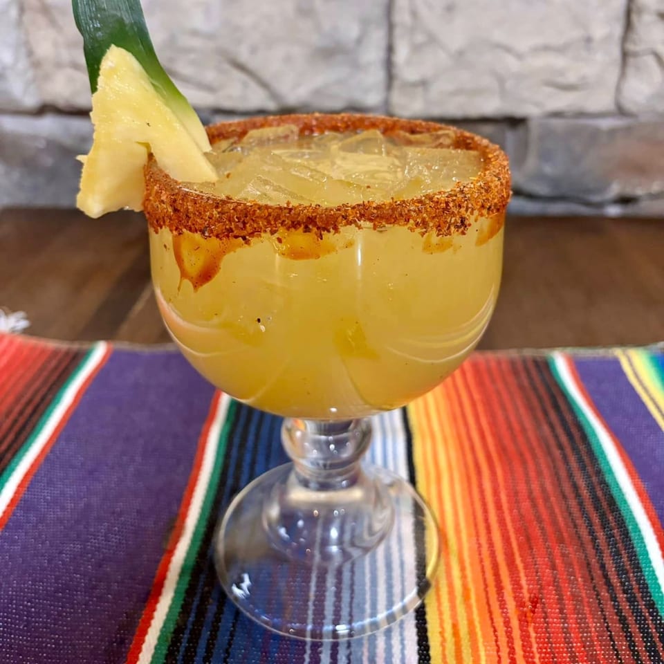 A pineapple margarita with a chili-lime rim and a pineapple wedge, on a colorful striped placemat.