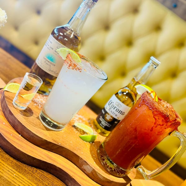 Mexican drinks on a wooden tray: a margarita, a shot of tequila, and a Michelada with a Corona.