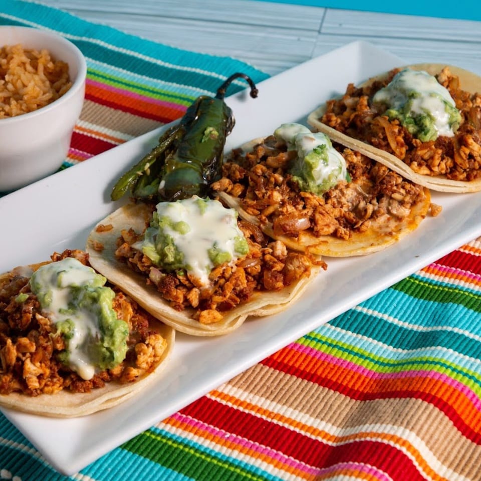 Four El Sazon Mexican Grill tacos with meat, guacamole, and cheese sauce, served with rice and a pepper.