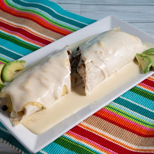 Two burritos covered in white cheese sauce, garnished with avocado slices on a white plate.