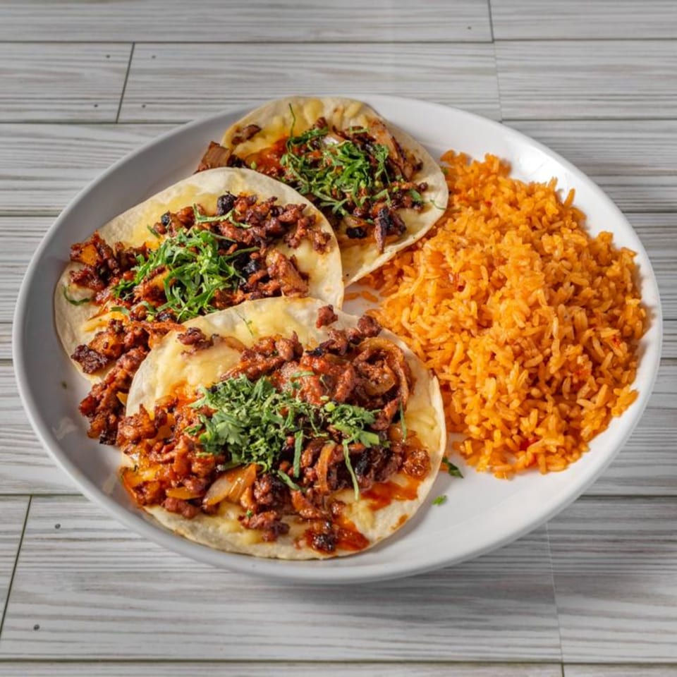 Three El Sazon Mexican Grill tacos with meat, cilantro, and rice on a white plate.