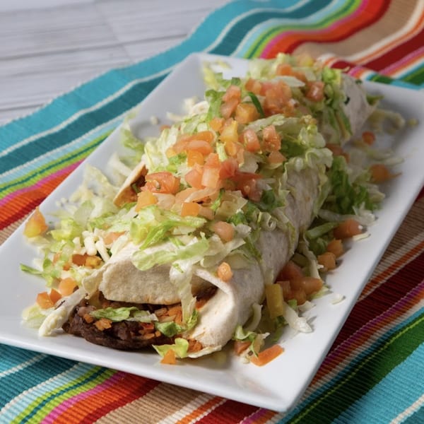 A large El Sazon Mexican Grill burrito, topped with shredded lettuce and diced tomatoes, on a white plate.