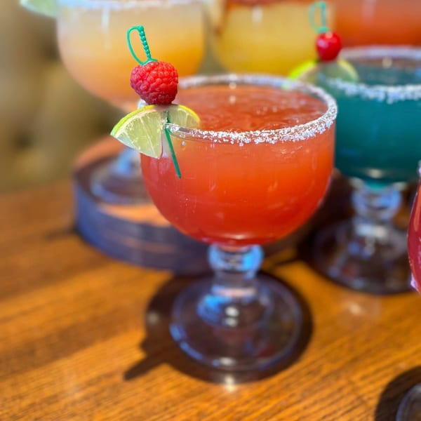 A vibrant red margarita with a salted rim, garnished with a lime wedge and raspberry, on a wooden table.